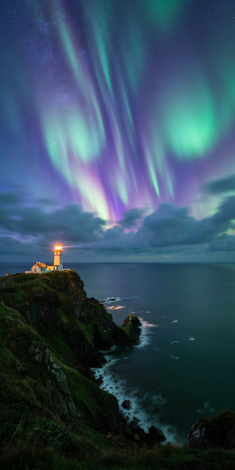 Photograph of a white lighthouse on a dark grassy sea cliff under swirling violet and teal aurora, its warm light glowing above the calm sea
